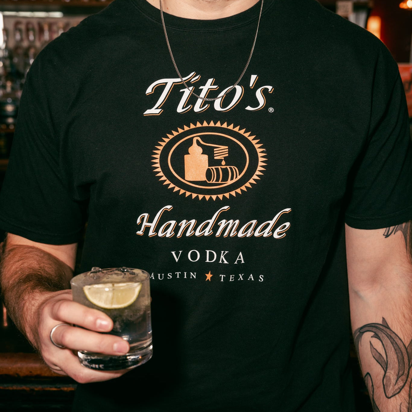 Person wearing a Classic Tito's Tee holding a Tito's Soda Lime cocktail in a bar setting.