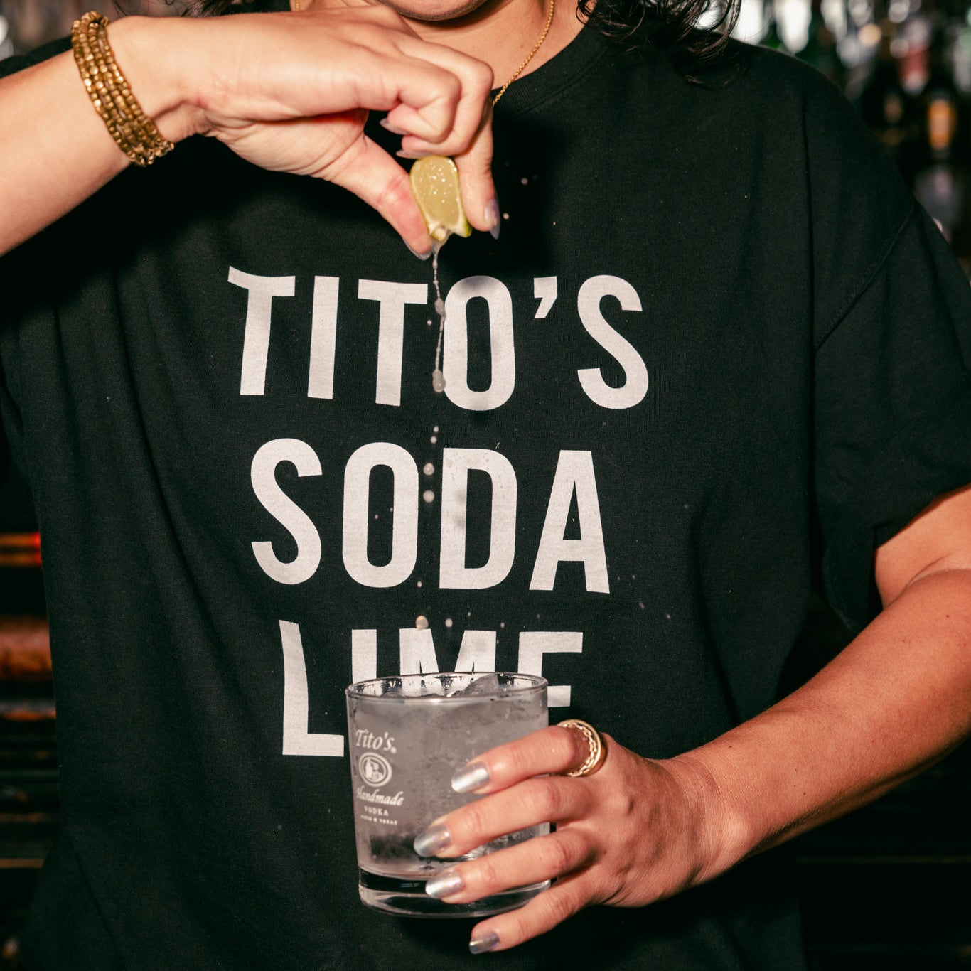 person wearing a Black Tito's Tee with a Twist of Lime squeezing a lime into a Tito's Soda cocktail