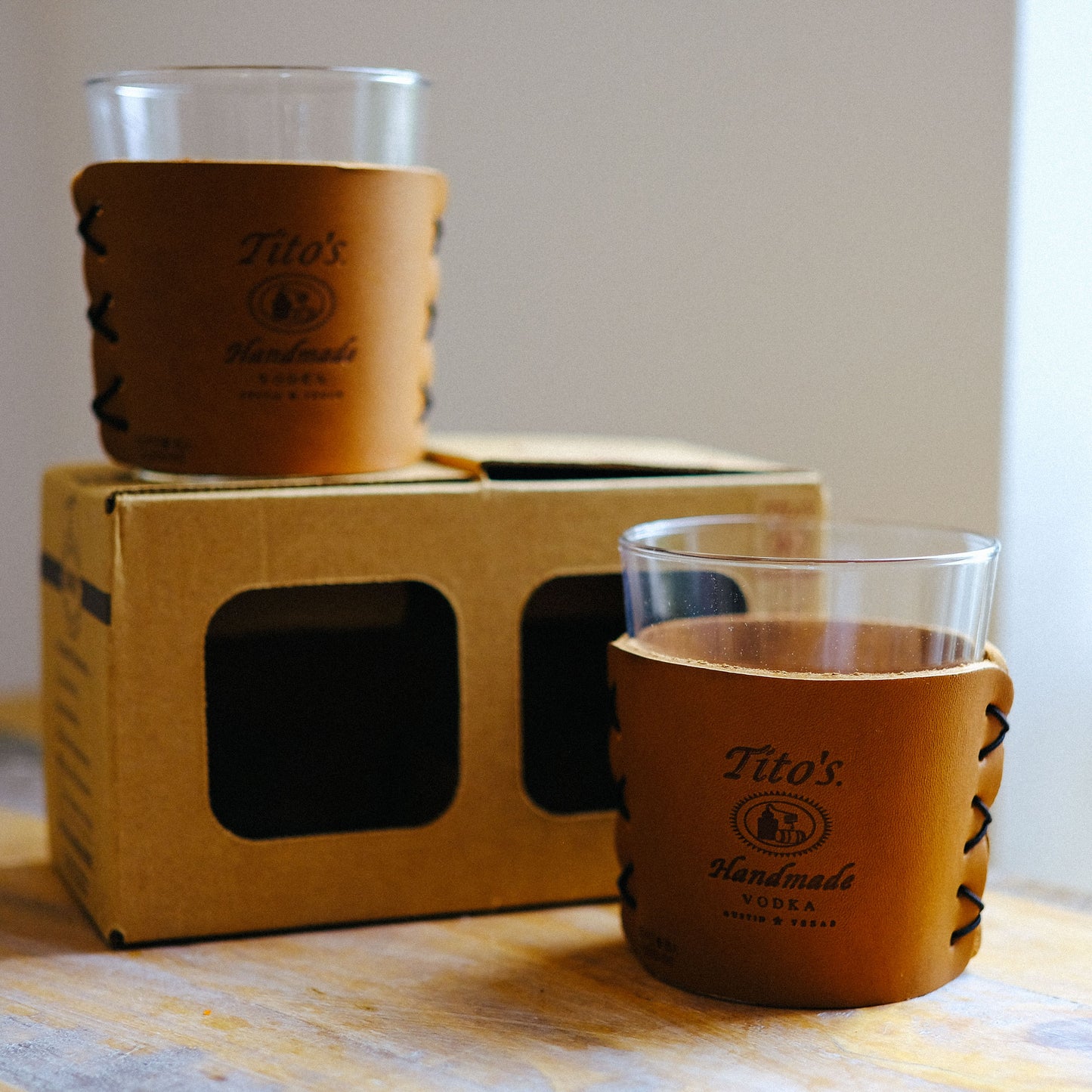 Two highball cocktail glasses in leather sleeves with Tito's Handmade Vodka logo on table