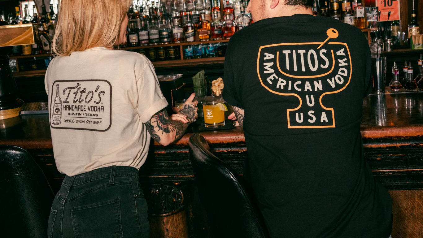 Two people at a bar wearing Tito's Classic Crewneck in Rust and Evergreen