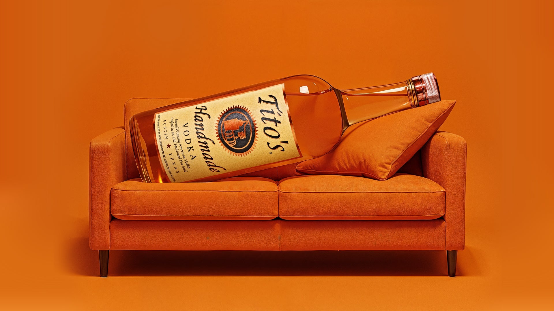 A Tito's Handmade Vodka bottle laying on an orange couch with an orange backgroun