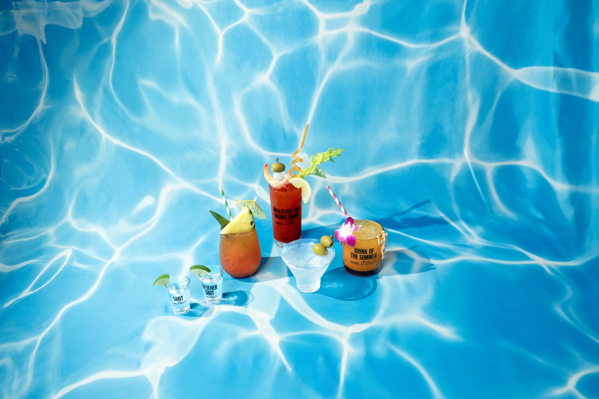 Clear and amber cocktail glasses filled with drinks, each glass printed with a different quirky phrase, background is blue and pool-esque