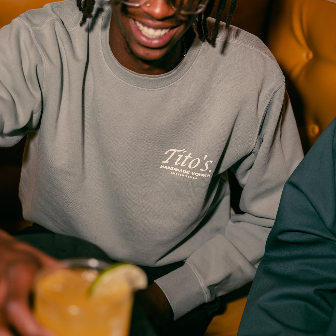 Front view of a person wearing the Tito's Austin Crewneck in a booth holding a Tito's cocktail