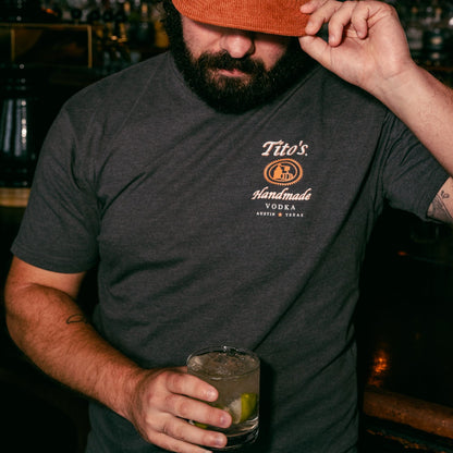 Front view of a person wearing the Tito's Austin Tee at a bar holding a Tito's cocktail