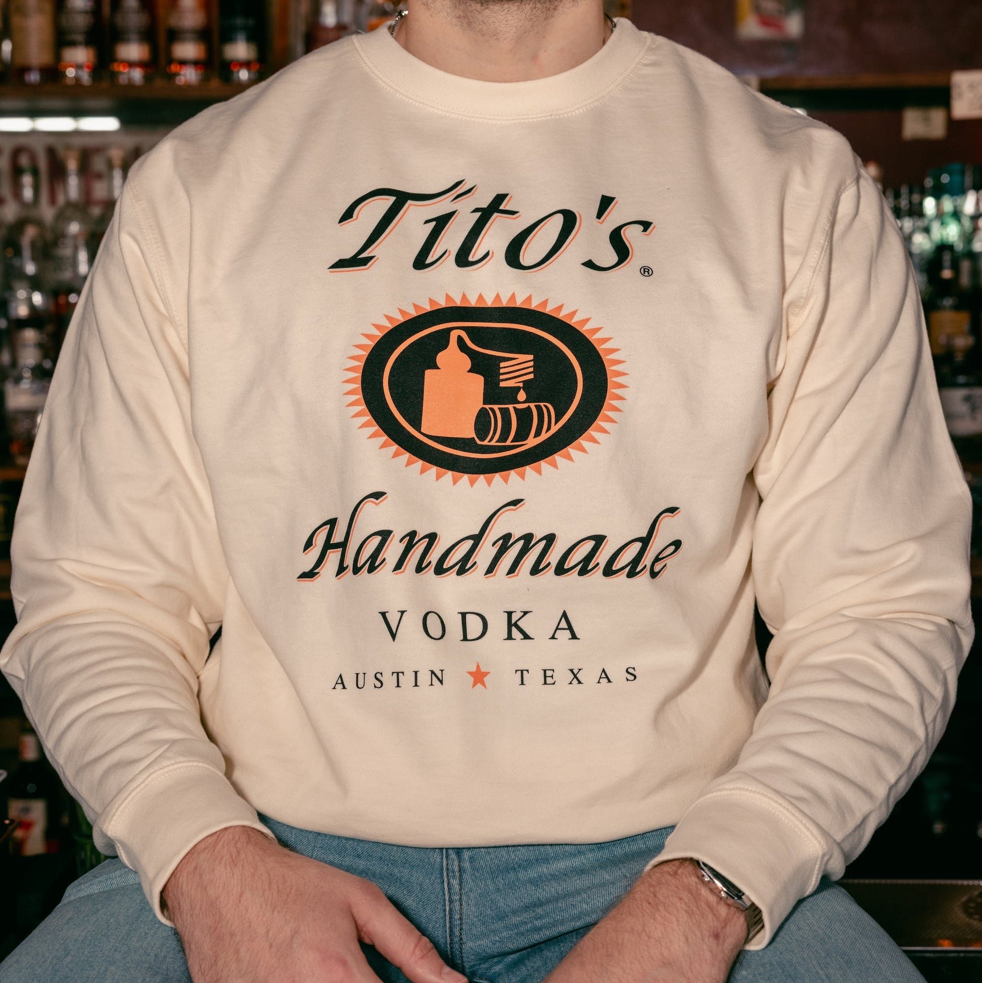 Person sitting on a bar wearing the cream colored Tito's Classic Crewneck