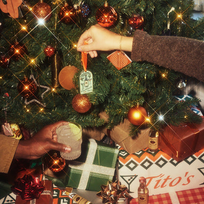 Person putting a Tito's Merry Martini Ornament on a Christmas tree and another person holding a cocktail