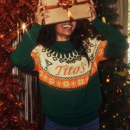 Person wearing the Tito's Ugly Sweater holding a gift box in front of a decorated Christmas tree.