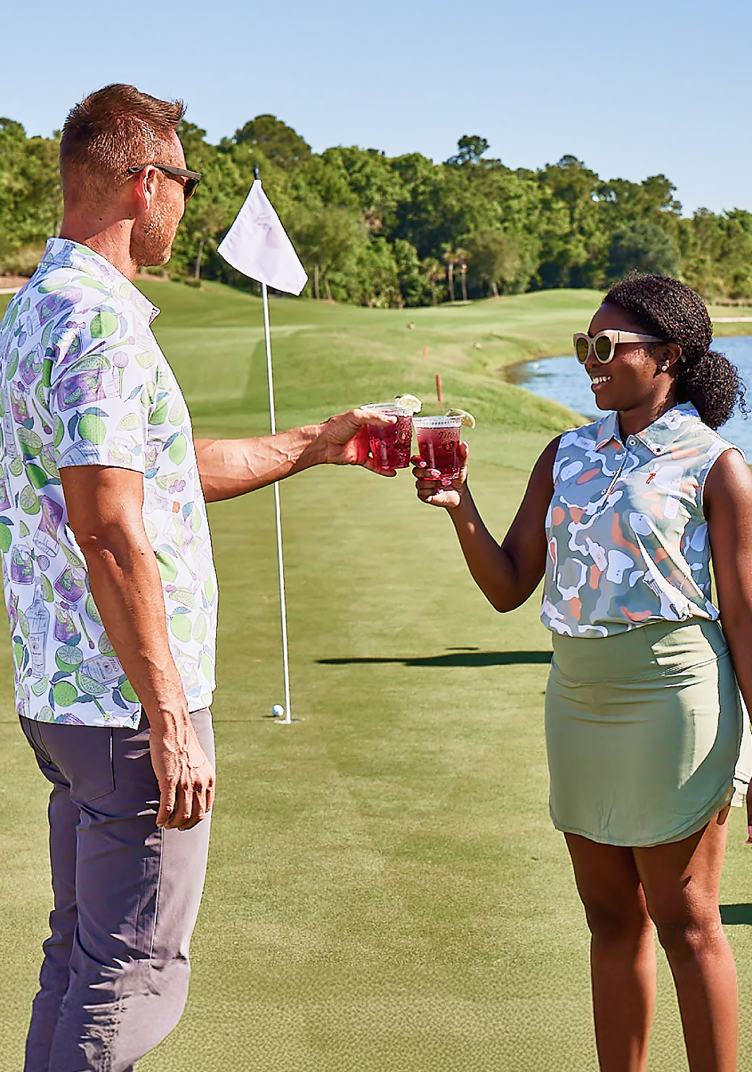A man standing on the left and a women standing on the right both wearing Tito's golf polo's, holding a Tito's Transfusion on a golf course.