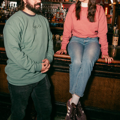 Two people sitting at a bar counter, one wearing the evergreen Tito's Classic Crewneck and the other wearing the rust colored Tito's Classic Crewneck