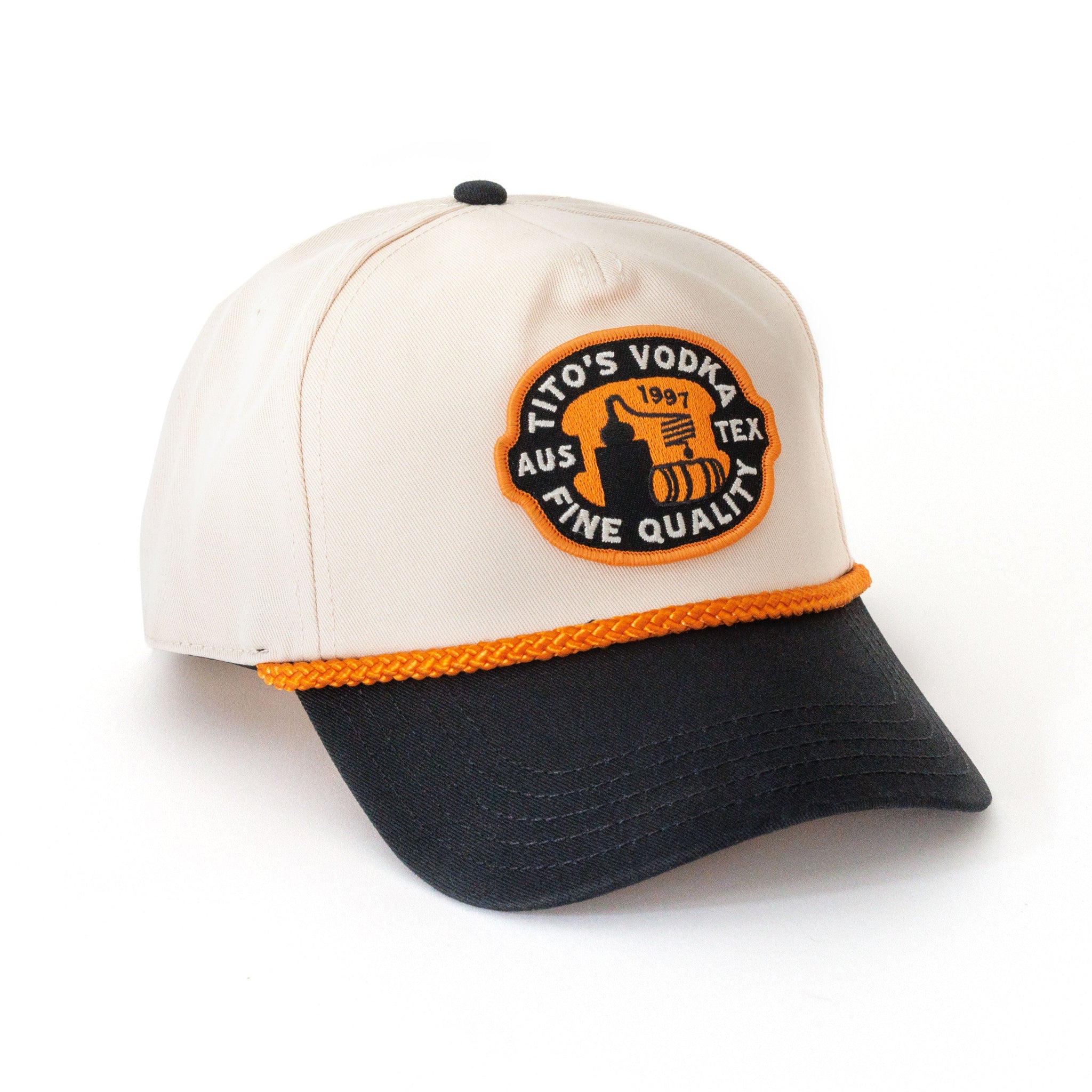 Front view of ivory hat with black brim, orange rope across seam, and a black, orange, and white patch with Tito's Vodka and a potstill. 