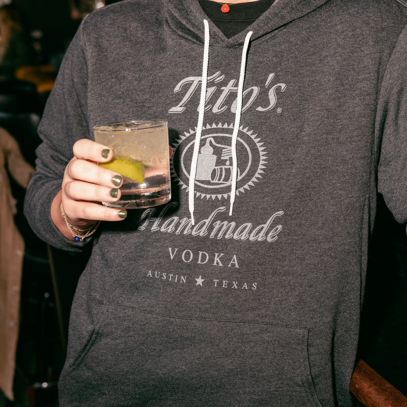 Person wearing a grey hoodie with Tito's Handmade Vodka logo, holding a Tito's Soda Lime cocktail.