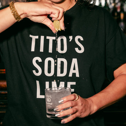 person wearing a Black Tito's Tee with a Twist of Lime squeezing a lime into a Tito's Soda cocktail 