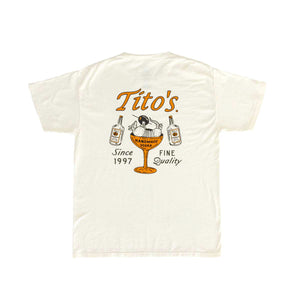 Back of cream short sleeve shirt with orange Tito's wordmark, 2 Tito's bottle, and an armadillo holding an olive in a martini glass, with "Since 1997" and "fine quality" text