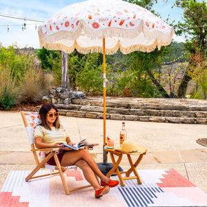 Woman sitting outside on the Tito's X Business & Pleasure Patio Chair under a Tito's X Business & Pleasure Patio Umbrella