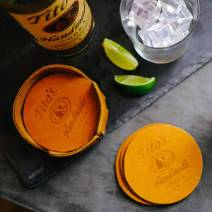 Set of leather coasters branded with Tito’s Handmade Vodka logo on a table next to leather coaster holder, limes, cocktail glass, and Tito's bottle