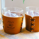 Detail view of highball cocktail glasses in leather sleeves with Tito’s Handmade Vodka logo and black stitching on a table