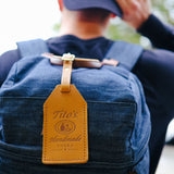 Front view of brown leather luggage tag with Tito’s Handmade Vodka logo attached to a backpack