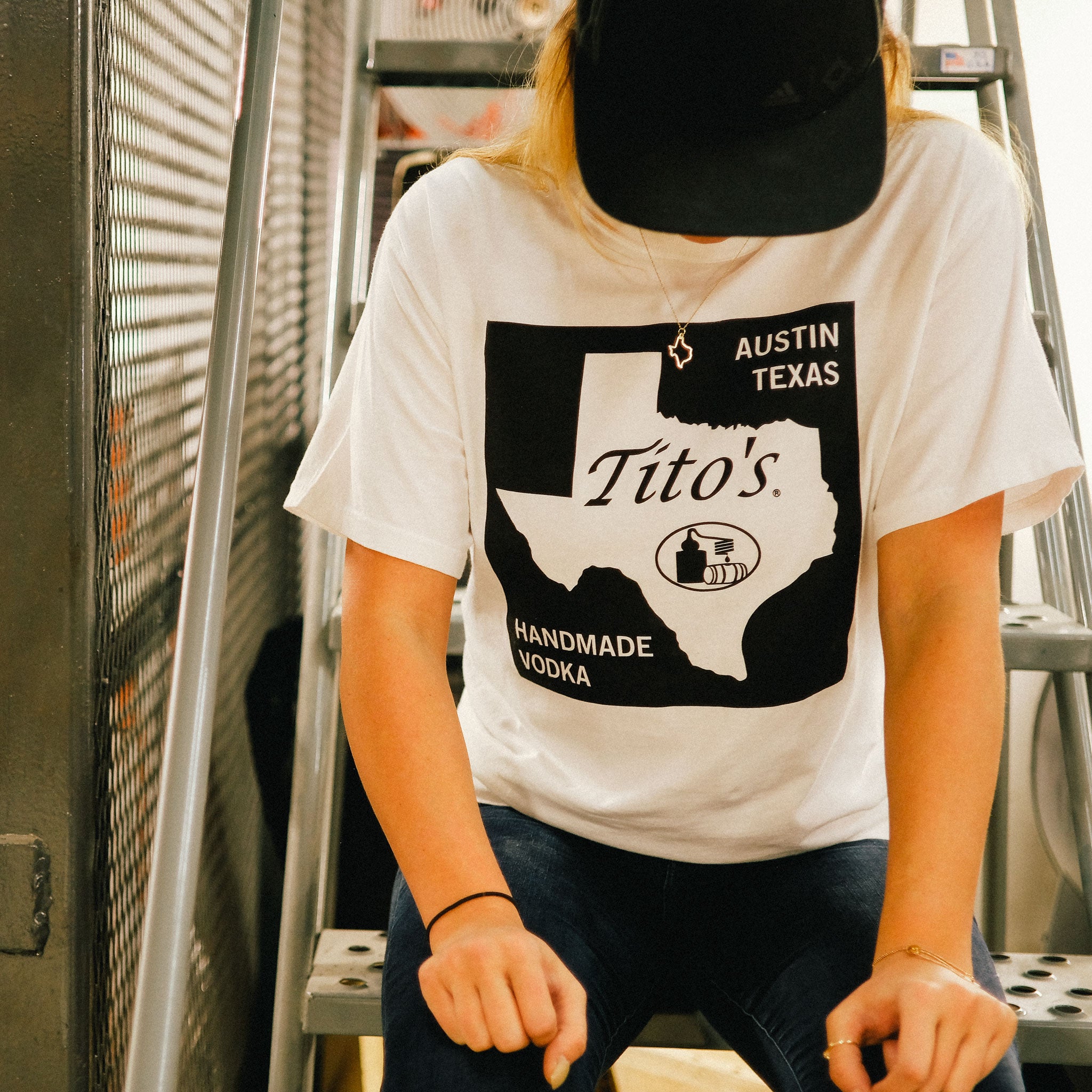 Girl sitting on stairs wearing white short-sleeve t-shirt featuring Texas state road sign design with Tito's Handmade Vodka mark