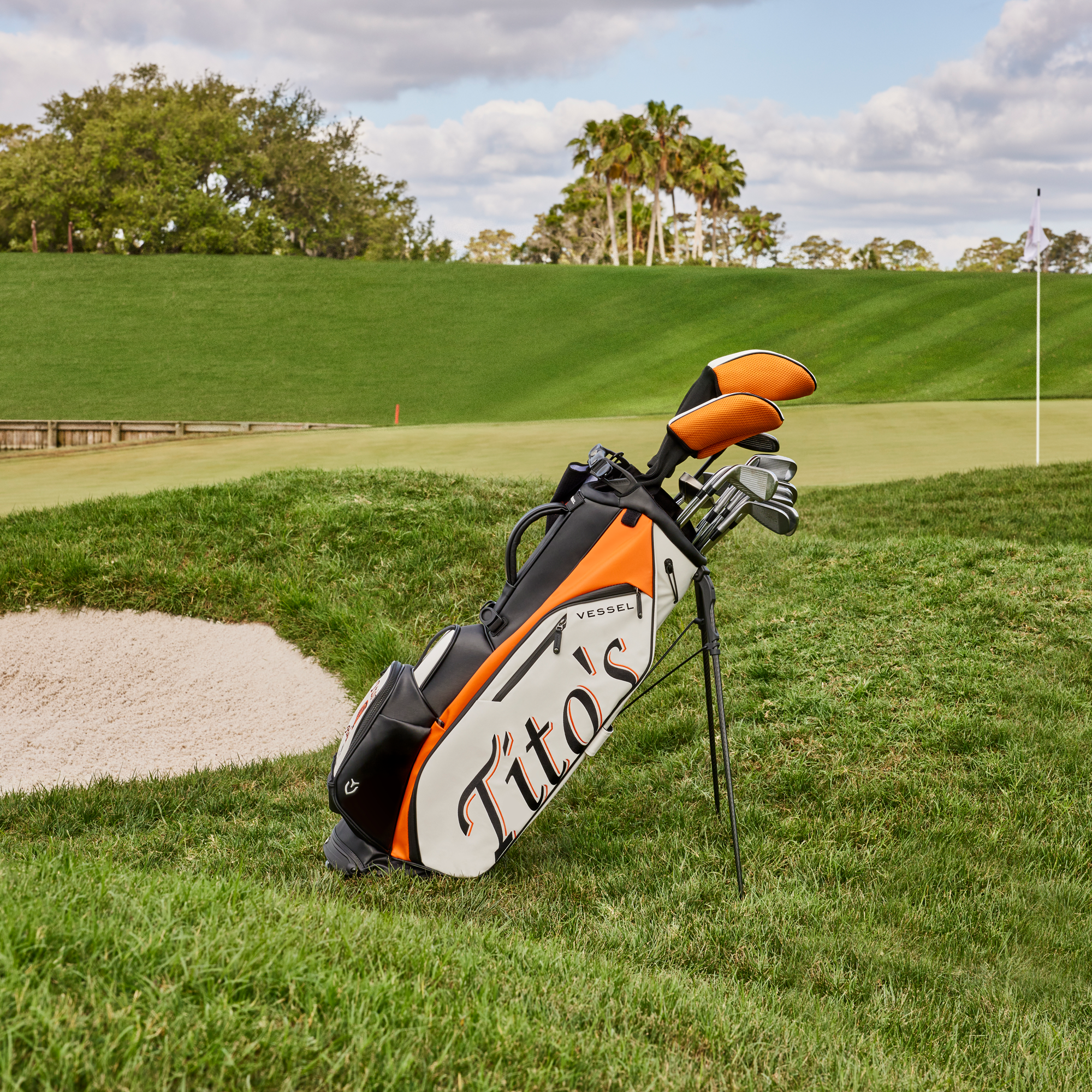 Side of white, orange, and black golf bag on a golf course with Tito's wordmark and VESSEL logo 