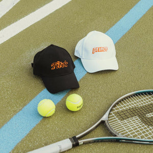 black hat with tito's logo and white hat with prince logo on a tennis court