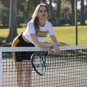 Woman leaning on tennis net wearing white crop top and black shorts