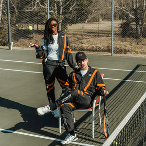 Man and woman on a tennis court wearing black track jacket and black windbreaker pants